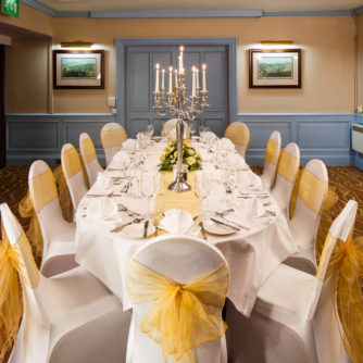 The Gillian Suite at Mercure Livingston Hotel, set up for a wedding breakfast, pretty blue painted wood panelling, yellow sashes, white linen, silver candlestick
