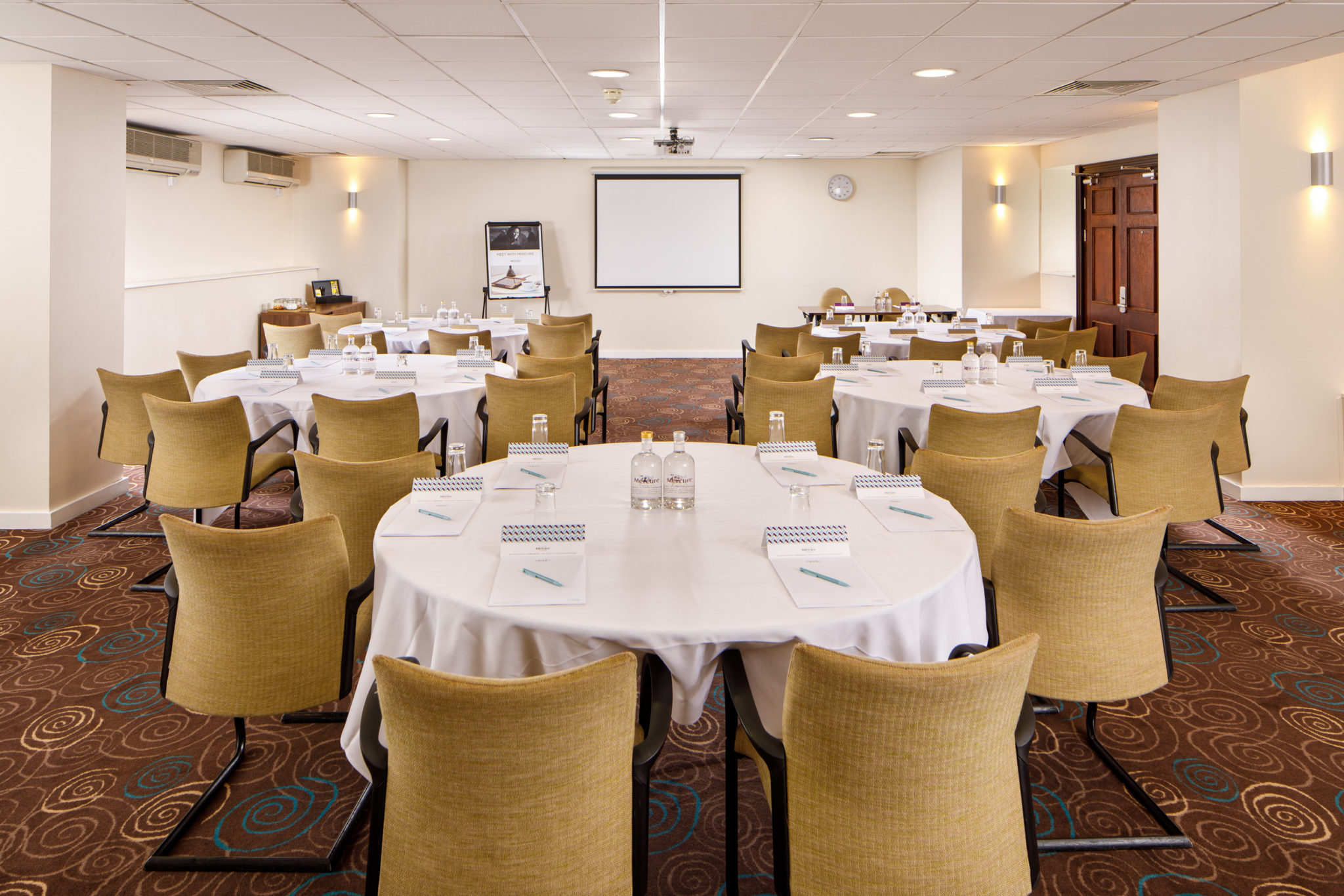 Mercure-Manchester-Piccadilly-Hotel-Portland-Meeting-Room-05-lr-080320 Long table seating 18 in The Congress Room at mercure gloucester bowden hall hotel ready for a meeting, with notepads, bottles of water and bowls of apples on the table
